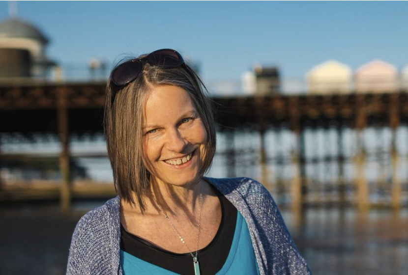 Nicky smiling on a beach, with a pier and colourful beach huts behind her